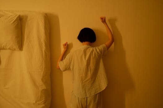 A person stands facing a wall with fists raised, symbolizing frustration or despair in a dimly lit room.